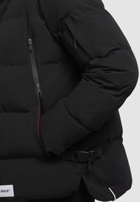 Jachetă puffer neagră cu un material exterior lucios, cusături vizibile, fermoar pe lateral și detaliu cu curea ajustabilă la talie.