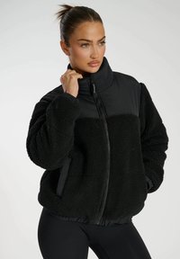Schwarze Jacke aus einem strukturierten, fleeceartigen Material, mit einem glatten schwarzen oberen Teil, Reißverschluss und elastischen Bündchen sowie einer taillierten Silhouette.
