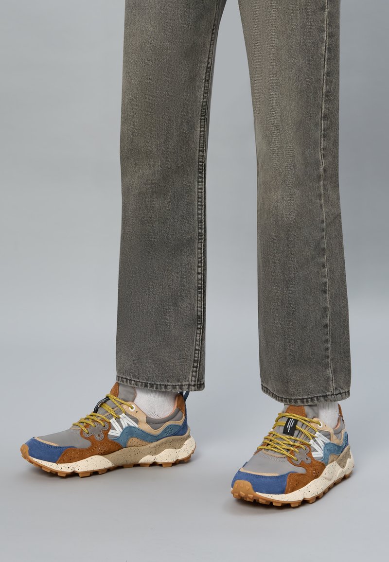 Večbarvni supergi z sivo, modro in rjavo sintetično materialom, teksturiranim podplatom in rumenimi vezalkami, nošeni z sivimi ozkimi kavbojkami.