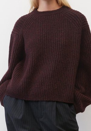 Femme portant un pull en maille côtelée marron foncé associé à un pantalon à fines rayures foncées, les mains dans les poches.