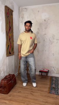 Homme avec des dreadlocks portant un t-shirt beige et un jean gris se tient dans une pièce aux murs en plâtre usés, sol en bois, sac en cuir et tenture murale à motifs.