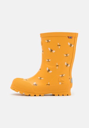 Botte de pluie en caoutchouc jaune avec des motifs d'abeilles noir et blanc, semelle et bordures texturées, et une petite étiquette en tissu rouge, blanc et bleu près du haut.