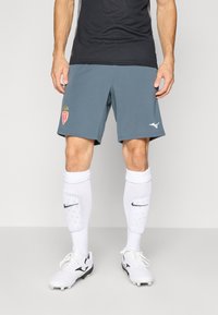 Grijze sportshorts voor heren met logo, gecombineerd met witte kniekousen met een zwart logo, en witte voetbalschoenen met zwarte accenten.