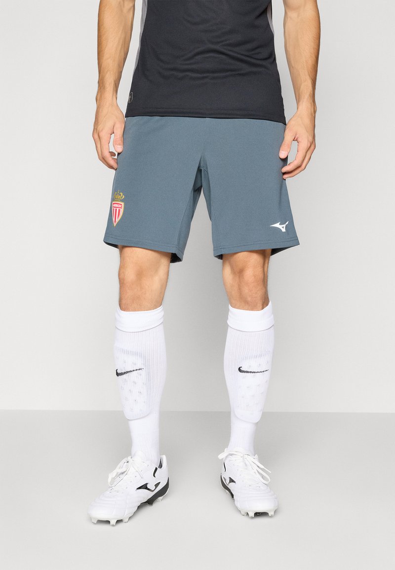 Grijze sportshorts voor heren met logo, gecombineerd met witte kniekousen met een zwart logo, en witte voetbalschoenen met zwarte accenten.