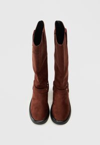 Botas altas de ante marrón con una textura suave, suelas negras planas y detalles en forma de círculo en el tobillo. Abiertas en la parte superior.