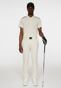 Ljust beige golfoutfit bestående av en polo med krage och matchande byxor, samt en svart bälte och handskar, och en svart keps.
