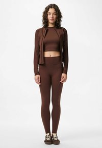 Jeune femme debout, portant un crop top à manches longues marron, des leggings assortis et des baskets marron avec des lacets clairs, sur un fond blanc uni.