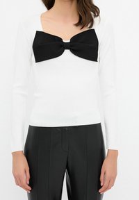 Haut blanc à manches longues avec un profond décolleté carré, orné d'un grand nœud noir sur la poitrine, associé à un pantalon noir ajusté.