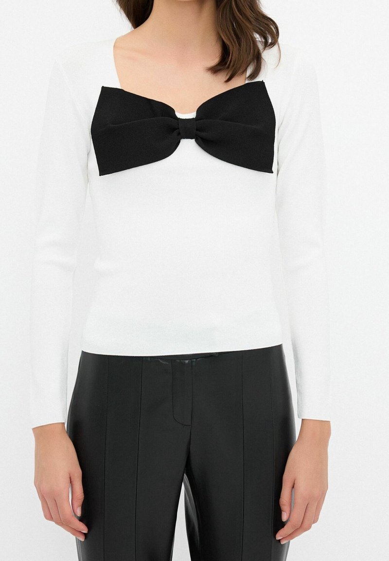 Haut blanc à manches longues avec un profond décolleté carré, orné d'un grand nœud noir sur la poitrine, associé à un pantalon noir ajusté.