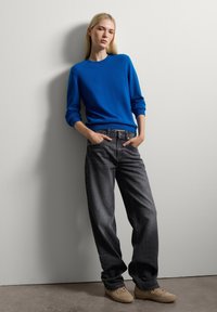 Suéter azul de manga larga combinado con jeans de tiro alto en gris oscuro. Zapatos de color claro. La modelo posa con las manos en los bolsillos junto a una pared.