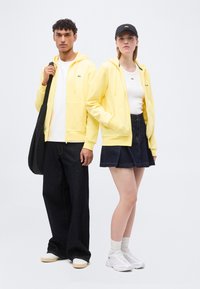 Homme et femme portant des sweats à capuche jaunes assortis, hauts blancs ; homme avec sac noir et pantalon large noir, femme avec casquette noire, jupe et baskets blanches.