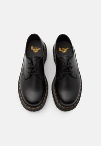 Chaussures basses Dr. Martens en cuir noir avec coutures jaunes et lacets noirs sur fond blanc.