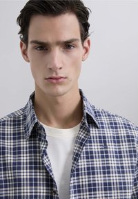 Camisa de cuadros en azul marino y blanco con bolsillo en el pecho, sobre una camiseta blanca lisa. Textura suave y detalle del collar visibles.
