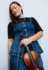Jeansvest met knopen, gedragen over een zwart shirt, met een bijpassende denim rok; houdt een houten viool in één hand.