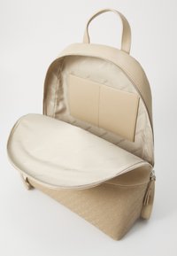 Zaino beige con un esterno in morbida pelle sintetica, chiusura con zip, foderatura in tessuto e una tasca interna. Presenta un dettaglio del logo testurizzato alla base.