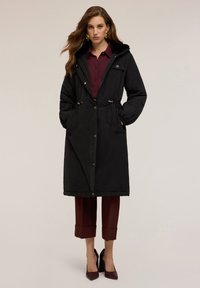 Svart vadderad lång kappa med huva, dragsko i midjan, framfickor och tryckknappar, kombinerad med en vinröd skjorta och bruna korta byxor.