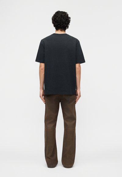 Άνδρας με κατσαρά μαλλιά που φοράει απλό μαύρο κοντομάνικο μπλουζάκι και καφέ παντελόνι, στέκεται με το πρόσωπο προς ένα λευκό φόντο.