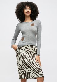 Femeie cu păr creț, purtând un top cu mâneci lungi gri cu imprimeuri florale maro și o fustă cu imprimeu zebră maro și crem, pozând cu o mână în buzunar.