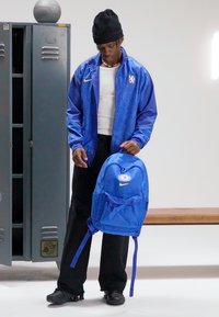 Kék Nike hátizsák logóval, nejlonból készült, állítható pántokkal és első zsebbel. Egy kék kabátot és fekete nadrágot viselő személy viseli.