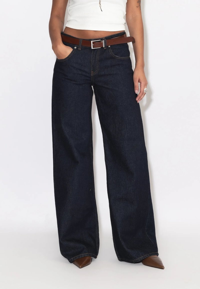 Person iført mørke vidde jeans med brun bælte, hvid cropped top og brune spidse sko, stående mod en hvid baggrund.