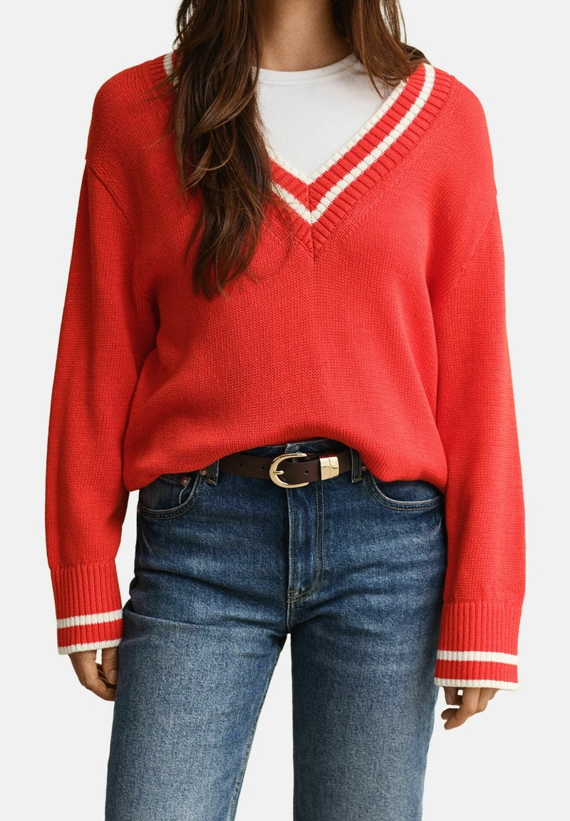 Femme portant un pull en maille rouge à col en V avec des rayures blanches sur le col et les poignets, rentré dans un jean bleu avec une ceinture marron.