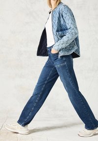 Quiltad denimjacka i ljusblå med diamantmönster, tillsammans med mörkblå jeans och beige sneakers, på en strukturerad neutral bakgrund.