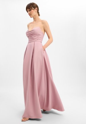 Vrouw in een strapless, vloerlang stofroze jurk met een nauwsluitend lijfje en geplooide rok, staand met één hand in haar zak.