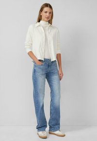 Biała koszula zapinana na guziki na kremowym golfie, zestawiona z jasnoniebieskimi dżinsami i beżowymi trampkami. Gładkie tkaniny i luźny krój.