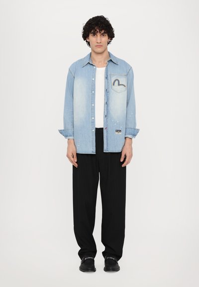 Camicia in denim, azzurra chiara con zone sbiadite, maniche lunghe, stile a bottoni, tasca frontale con un design arcobaleno, abbinata a pantaloni neri a righe.