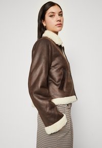 Giacca di pelle marrone con colletto e polsini in shearling, con chiusura a zip sul davanti e una tasca laterale, abbinata a una gonna a effetto houndstooth.