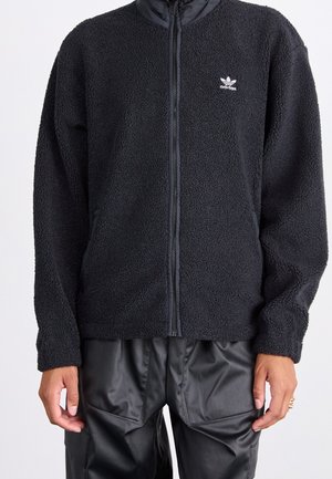 Sort fleece-lynlåsjakke med Adidas-logo på venstre bryst, båret med sorte læder-look bukser, vist fra hals til hofter.