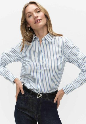 Femme portant une chemise blanche à rayures verticales bleues, un jean foncé et une ceinture noire avec une boucle argentée, posant les mains sur les hanches.