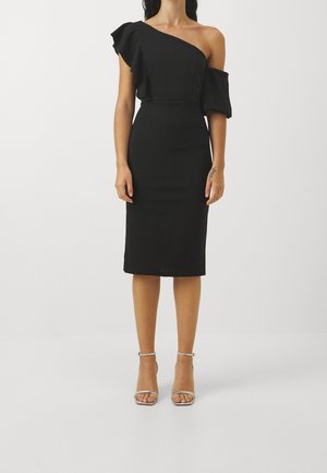 Frau trägt ein schwarzes, asymmetrisches Midikleid mit Rüschen am Ärmel und silbernen High-Heel-Sandalen, steht vor einem weißen Hintergrund.