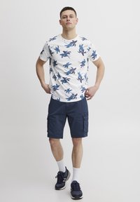 Tricou alb cu imprimeu floral bleu, asortat cu pantaloni scurți cargo bleumarin. Modelul poartă șosete albe și teniși bleumarin. O ținută simplă și lejeră.