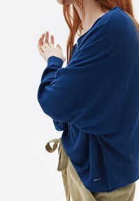 Blauwe, lange mouwen trui met een ontspannen pasvorm en geribbelde manchetten, gecombineerd met beige broeken met een gestrikt taille. Zachte gebreide textuur, minimale details.