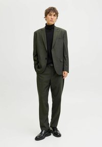 Completo verde oliva con giacca aderente e pantaloni a pieghe, abbinato a un dolcevita nero. Scarpe nere e dettagli hardware minimal.