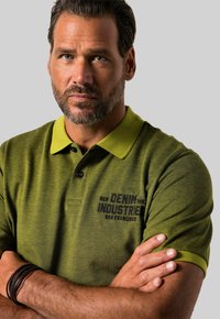 Vihreä polo-paita, jossa on erottuva keltainen kaulus, teksturoitu kangas ja brodeerattu "DENIM INDUSTRIES SAN FRANCISCO" -logo.