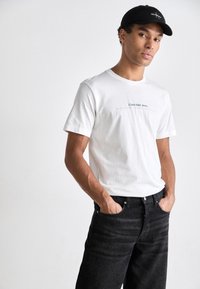 Weißes Baumwoll-T-Shirt mit Rundhalsausschnitt und schwarzem "Calvin Klein Jeans"-Logo. Kombiniert mit dunkelblauen Jeans und einer schwarzen Kappe. Hände in den Taschen.
