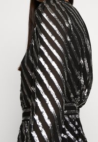 Robe noire transparente avec des rayures métalliques argentées, encolure ronde, manches bouffantes et taille cintrée. Le tissu texturé ajoute un intérêt visuel.