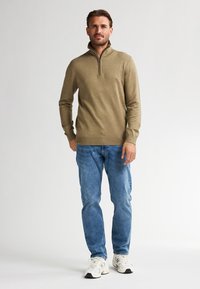 Beige Zip-Up-Pullover mit hohem Kragen, kombiniert mit hellblauen Jeans und weißen Sportschuhen. Glatte Textur, lockere Passform und lange Ärmel.