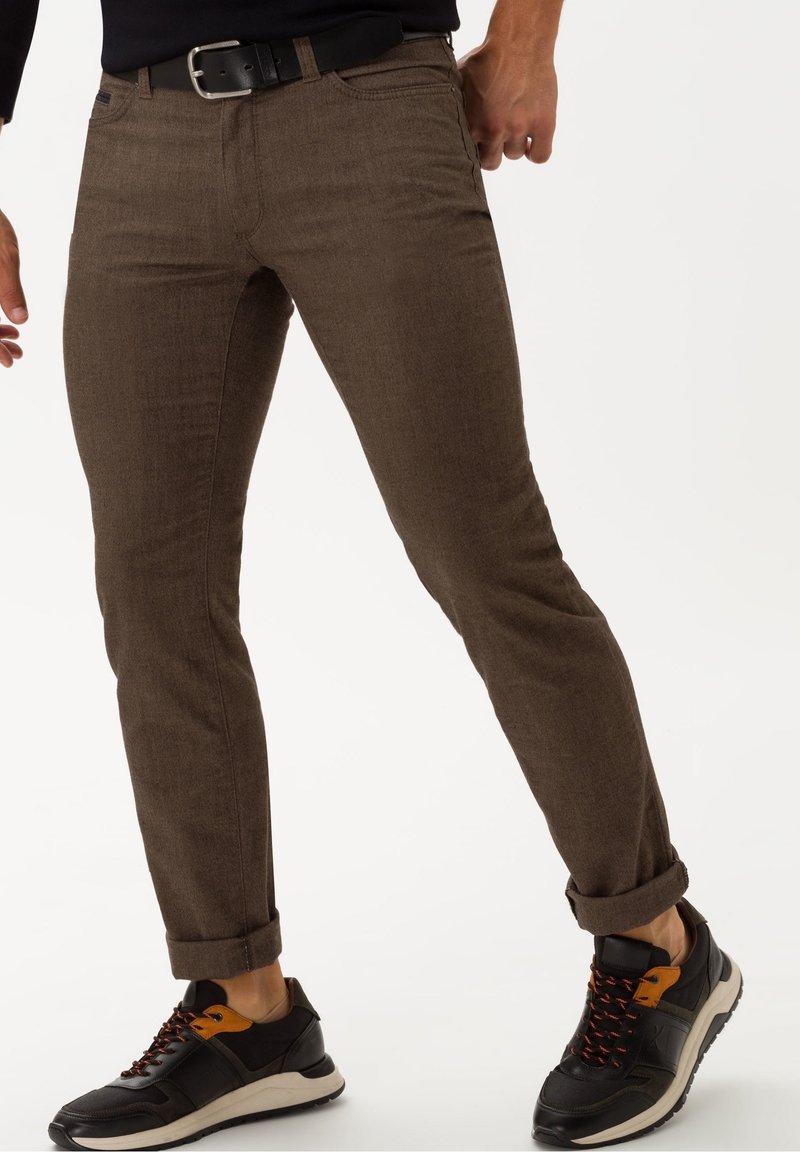Brown, slim-fit trousers with a smooth texture and rolled cuffs, paired with black and orange sneakers. Metal belt buckle visible.