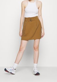 Falda marrón ajustada con un cinturón y hebilla circular, con una cintura alta y una textura suave, combinada con una blusa blanca y zapatillas coloridas.