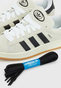 Baskets Adidas Campus beiges avec des rayures noires et des lacets blancs, accompagnées d'une paire de lacets noirs Adidas avec une étiquette bleue de marque.