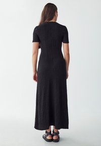 Schwarz, strukturiertes Maxikleid mit kurzen Ärmeln und tailliertem Schnitt, mit einem Rückenverschluss und kombiniert mit schwarzen Sandalen.