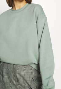 Felpa oversize verde chiaro con scollo rotondo e maniche lunghe, abbinata a una gonna plissettata grigia a quadri. Tessuto morbido in misto cotone.