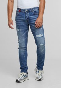 Kék kopott farmernadrág, slim fit szabással, szakadt és szétfont részletekkel. Gombos sliccsel és egy piros címkével a derékrészen.