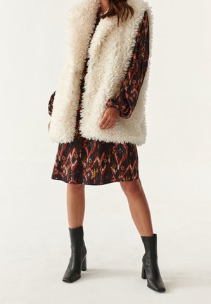 Femme portant des bottines noires à talons, un gilet sans manches blanc et duveteux, ainsi qu'une robe à manches longues à motifs dans des tons rouges et orange.