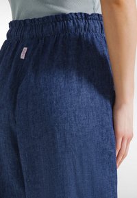 Zbliżenie osoby noszącej luźne ciemnoniebieskie spodnie dżinsowe z elastycznym pasem i małą białą metką marki po boku.