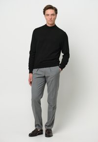 Svart långärmad tröja, grå plisserade byxor och mörka loafers. Tröjan har en ribbad halsringning; byxorna har ett subtilt mönster.