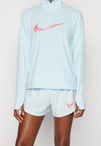 Jasnoniebieski sportowy pulower z ćwierć zamkiem i długimi rękawami, z różowym logiem Nike. Połączony z dopasowanymi jasnoniebieskimi szortami.